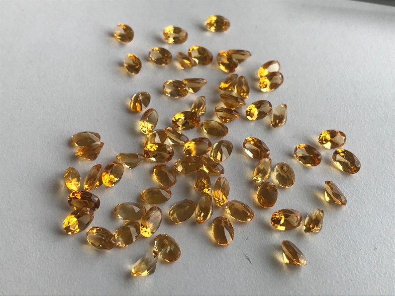May include: A collection of oval-shaped, faceted, golden-yellow gemstones. The gemstones are scattered on a white surface.