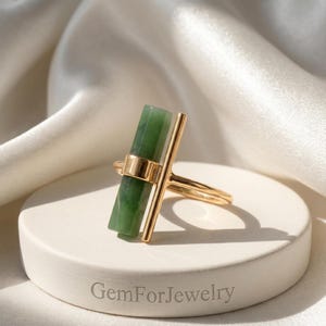 May include: A gold-colored ring featuring a rectangular, green gemstone. The ring has a vertical bar design and a band that wraps around the finger. The ring is displayed on a white, circular pedestal with the text "GemForJewelry".