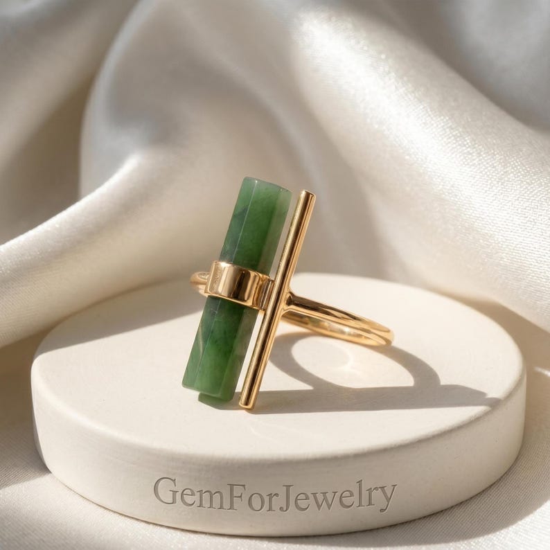 May include: A gold-colored ring featuring a rectangular, green gemstone. The ring has a vertical bar design and a band that wraps around the finger. The ring is displayed on a white, circular pedestal with the text "GemForJewelry".