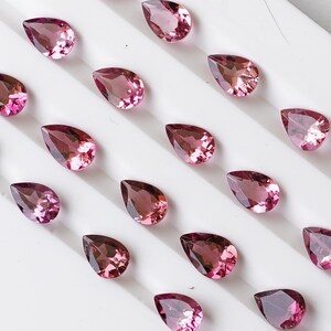 May include: A collection of pink pear-shaped gemstones arranged on a white surface. The gemstones are various sizes and have a subtle sparkle.