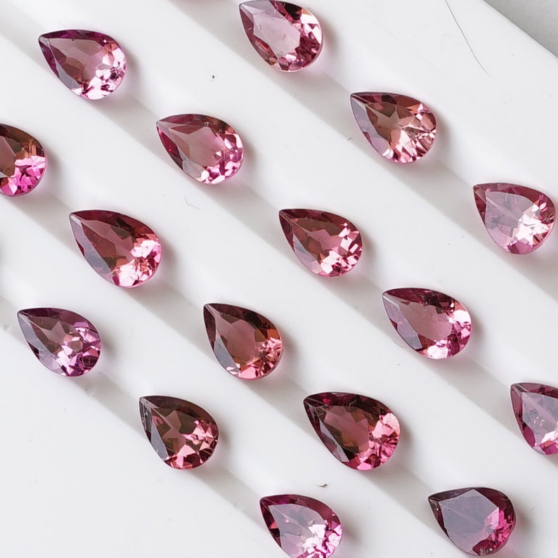 May include: A collection of pink pear-shaped gemstones arranged on a white surface. The gemstones are various sizes and have a subtle sparkle.