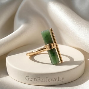 May include: A gold ring featuring a rectangular green gemstone and a vertical gold bar. The ring is set on a white, circular pedestal with the text "GemForJewelry" engraved on the base. The background is a white, silky fabric.