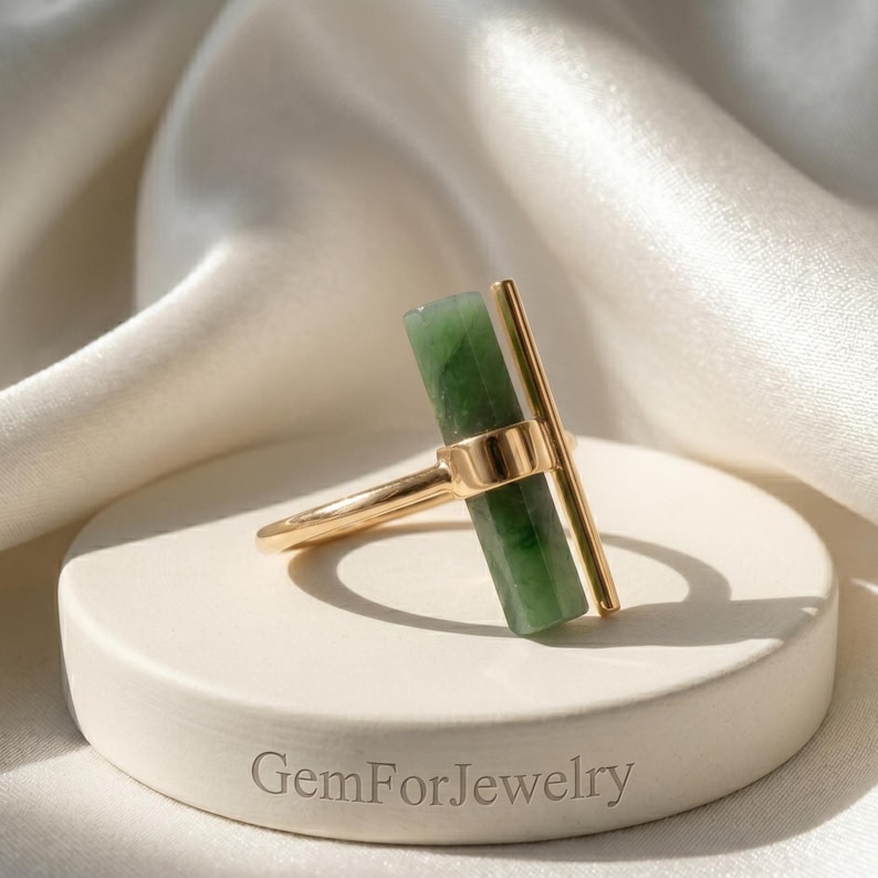May include: A gold ring featuring a rectangular green gemstone and a vertical gold bar. The ring is set on a white, circular pedestal with the text "GemForJewelry" engraved on the base. The background is a white, silky fabric.
