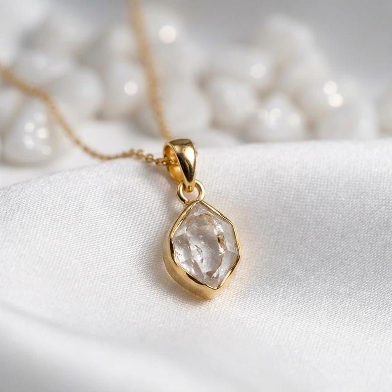 K&ouml;nnte beinhalten: Eine goldfarbene Halskette mit einem Anh&auml;nger aus klarem, facettiertem Edelstein. Der Anh&auml;nger hat eine einzigartige, unregelm&auml;&szlig;ige Form und ist in eine goldene L&uuml;nette gefasst. Die Halskette wird auf einer wei&szlig;en, strukturierten Oberfl&auml;che pr&auml;sentiert.