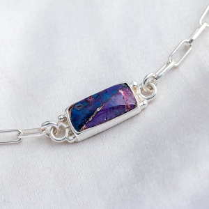 May include: A silver chain necklace featuring a rectangular pendant with a vibrant, multi-colored stone. The stone displays shades of blue, purple, and pink. The necklace is set against a white background, highlighting the jewelry's design.