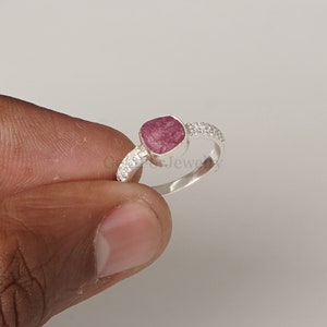Peut inclure: Une bague en argent avec une pierre pr&eacute;cieuse rose sertie dans une forme carr&eacute;e. La bague a une bande de petites pierres blanches.