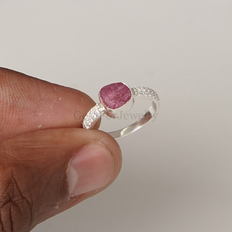 Peut inclure: Une bague en argent avec une pierre pr&eacute;cieuse rose sertie dans une forme carr&eacute;e. La bague a une bande de petites pierres blanches.