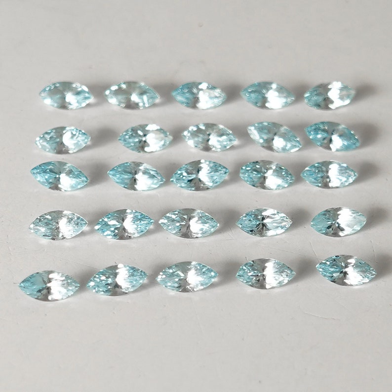 May include: Twenty-four marquise-shaped gemstones in a light blue color. The gemstones are arranged in four rows of six, on a white surface.