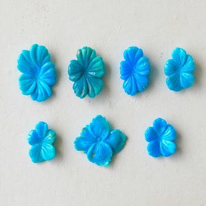 May include: Seven turquoise blue flower-shaped cabochons, each with a slightly different design, are arranged on a white surface.