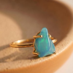 May include: A gold ring featuring a single, irregular, polished blue opal gemstone set in a four-prong setting. The opal displays internal flashes of green and blue light, and the ring band is a smooth, thin, polished gold metal wire.