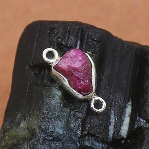 May include: A silver connector with a raw pink ruby gemstone. The connector has two loops on either side for attaching to jewelry.