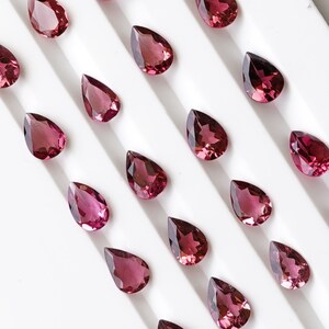 May include: A collection of pear-shaped gemstones arranged in rows on a white surface. The gemstones are a deep reddish-pink color and have a glossy finish.
