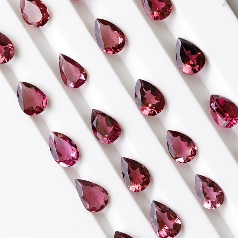 May include: A collection of pear-shaped gemstones arranged in rows on a white surface. The gemstones are a deep reddish-pink color and have a glossy finish.