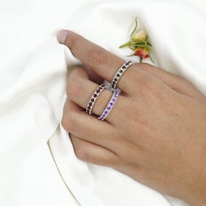 May include: Three silver rings with different colored gemstones. The rings are stacked on a finger. The top ring has purple gemstones, the middle ring has brown gemstones, and the bottom ring has clear gemstones.