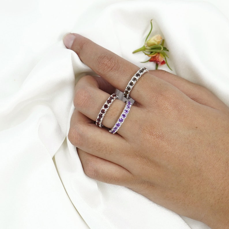May include: Three silver rings with different colored gemstones. The rings are stacked on a finger. The top ring has purple gemstones, the middle ring has brown gemstones, and the bottom ring has clear gemstones.