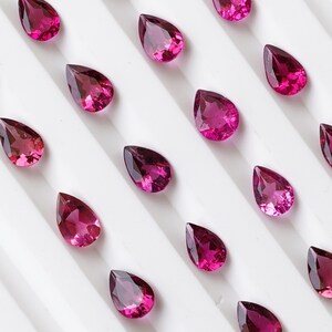 May include: A collection of pear-shaped pink gemstones arranged on a white surface. The gemstones are all the same size and shape, and they are arranged in a symmetrical pattern.