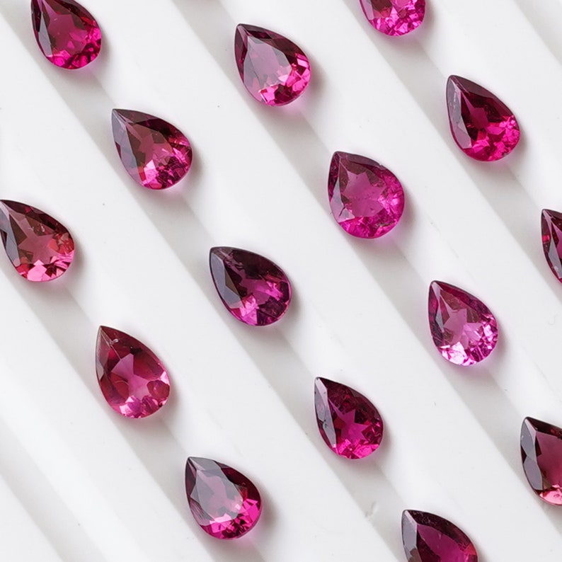 May include: A collection of pear-shaped pink gemstones arranged on a white surface. The gemstones are all the same size and shape, and they are arranged in a symmetrical pattern.