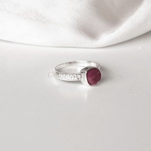Peut inclure: Une bague en argent avec une pierre pr&eacute;cieuse rouge sertie dans une forme carr&eacute;e. La bande de la bague est d&eacute;cor&eacute;e de petites pierres blanches.