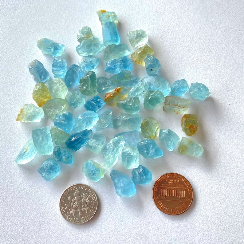 Pode incluir: Uma cole&ccedil;&atilde;o de pedras brutas de &aacute;gua-marinha azul claro, algumas com um tom amarelo, espalhadas sobre uma superf&iacute;cie branca. Um centavo americano &eacute; inclu&iacute;do para escala.