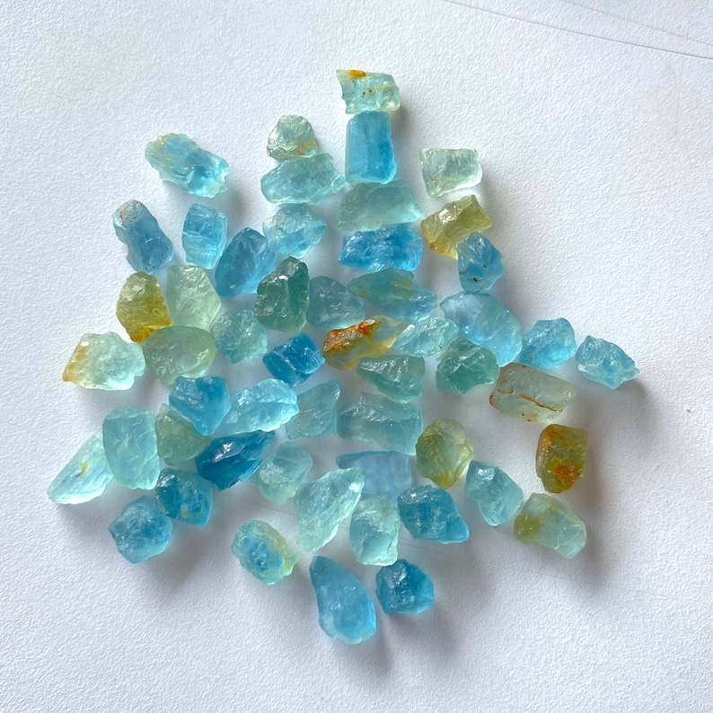 Pode incluir: Uma cole&ccedil;&atilde;o de pedras preciosas de &aacute;gua-marinha bruta, de cor azul claro com um tom amarelo-dourado. As pedras s&atilde;o de v&aacute;rios tamanhos e formas, e algumas t&ecirc;m um aspecto ligeiramente nublado.