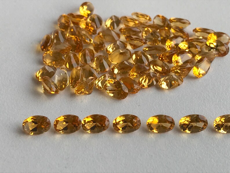 May include: A large pile of oval-shaped, golden yellow gemstones, with a smaller row of seven gemstones in the bottom of the image.