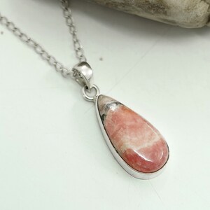 May include: A silver necklace featuring a teardrop-shaped pendant. The pendant has a pink and white stone with a silver bezel. The chain is a delicate silver link style. The necklace is displayed on a white surface.