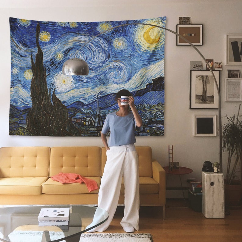 May include: A blue and yellow tapestry with a swirling starry night sky design. The tapestry is hanging on a white wall in a living room setting. A person is standing in front of the tapestry, holding a mug in front of their face.