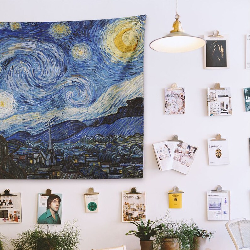 May include: A blue and yellow tapestry featuring a swirling night sky with stars and a crescent moon. The tapestry is hanging on a white wall with other framed artwork and a light fixture.