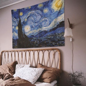 May include: A blue and yellow tapestry featuring a swirling night sky with stars and a crescent moon. The tapestry depicts a village with a church steeple in the distance. The tapestry is hanging on a wall above a bed with brown bedding.