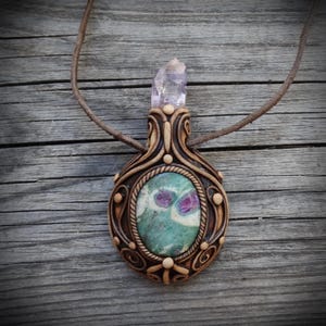 May include: A pendant necklace featuring a central oval-shaped green and magenta stone encased in a dark brown sculpted frame with swirling patterns and small round accents. A clear, pointed crystal sits at the top, attached to a brown cord.