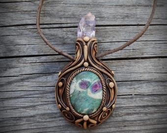 Puede incluir: Un collar con colgante que presenta una piedra central ovalada de color verde y magenta, engastada en un marco esculpido de color marrón oscuro con patrones de remolinos y pequeños detalles redondos. Un cristal transparente y puntiagudo corona la pieza, unido a un cordón marrón.