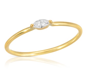 May include: A delicate gold ring featuring a marquise-cut diamond. The slender band is polished gold, with small gold accents on either side of the diamond. The ring is set against a white background, highlighting its elegant design.