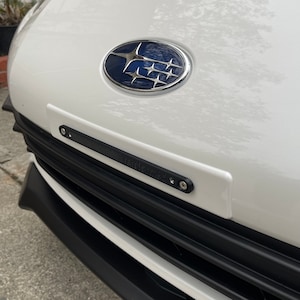 May include: The front of a white car with a black license plate holder. The holder is mounted below the Subaru emblem and above the black grill. The text "Vaughn3d" is visible on the holder.