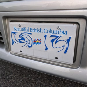 May include: A white license plate with blue text that reads "Beautiful British Columbia." The plate features a stylized design and the British Columbia flag. It is mounted on a silver car bumper.