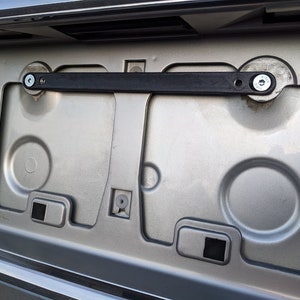 May include: A close-up of a car's license plate mounting area. The silver-colored area has various molded shapes and mounting points. A black bracket is secured with screws, and the overall design is functional.