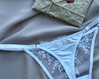 May include: White thong underwear featuring sheer mesh side panels with white floral embroidery, adjustable straps, and the gold embroidered date 05-08-25 on the front. Above the underwear sits a square brown paper envelope sealed with red wax.