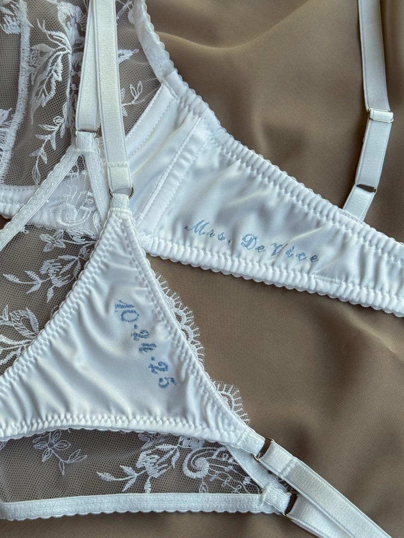May include: White lingerie set featuring delicate lace and satin. The bra and the bottom have blue embroidered text. The bra says "Mrs. De Vice" and the bottom says "10.24.25". The set is on a neutral background.