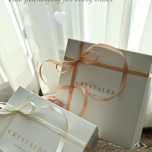 May include: Two white gift boxes with ribbons. The boxes have the text "CRYSTALBRA handmade lingerie" printed on them. The boxes are tied with light-colored ribbons. The image also includes the text "cute packaging for every order".