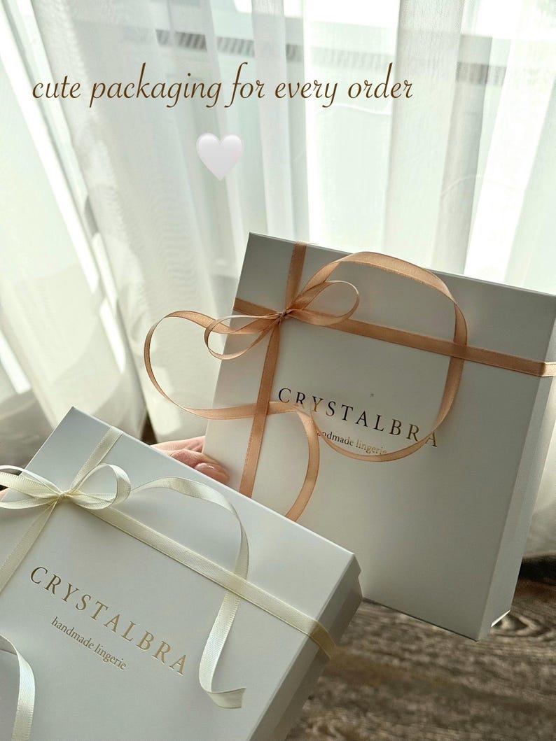 May include: Two white gift boxes with ribbons. The boxes have the text "CRYSTALBRA handmade lingerie" printed on them. The boxes are tied with light-colored ribbons. The image also includes the text "cute packaging for every order".