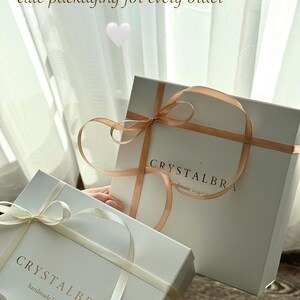 May include: Two white gift boxes with ribbons. The boxes have the text "CRYSTALBRA handmade lingerie" printed on them. The boxes are tied with light-colored ribbons. The image also includes the text "cute packaging for every order".