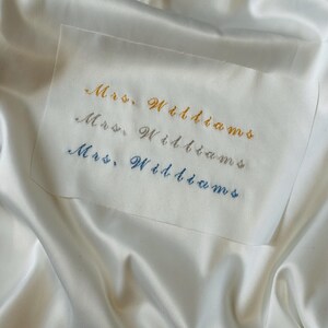 May include: A close-up of a white fabric with three lines of embroidered text that reads "Mrs. Williams" in gold, silver, and blue thread. The fabric has a satin-like sheen.