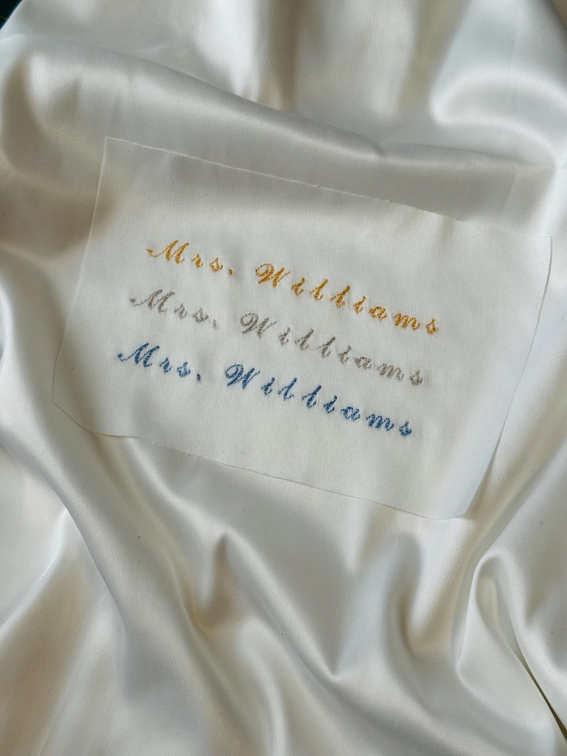 May include: A close-up of a white fabric with three lines of embroidered text that reads "Mrs. Williams" in gold, silver, and blue thread. The fabric has a satin-like sheen.