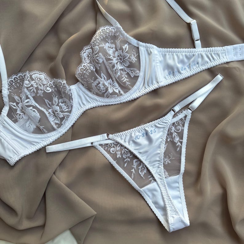 May include: White lingerie set featuring a bra and matching thong. The bra has floral lace detailing on the cups and straps. The thong also has floral lace accents. The set is made of a satin-like material.