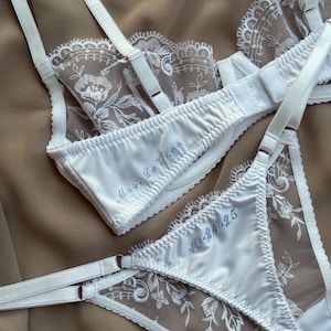 May include: White lingerie set featuring a bra and matching underwear. The bra and underwear are made of satin and lace. The bra has adjustable straps and the underwear has a delicate floral lace design. The bra is embroidered with "Mrs. De Vloe" and the underwear is embroidered with "10-24-25".