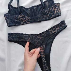 May include: A matching lingerie set featuring a bra and thong. The set is made of a sheer fabric with a leopard print pattern in shades of brown and black. The bra has adjustable straps and a hook-and-eye closure. The thong has a scalloped edge.