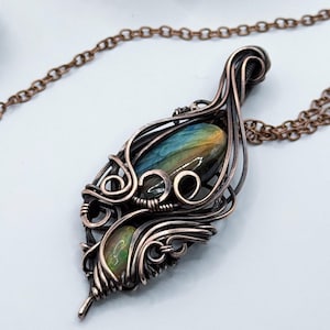 Può includere: Una collana con pendente artigianale in filo di rame. Il pendente presenta un design a goccia con un'intricata filigrana, che racchiude due pietre preziose lucide. Le pietre preziose mostrano sfumature di blu, verde e giallo. La collana include una catena di rame abbinata.