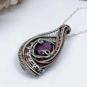 Può includere: Un ciondolo a forma di goccia con una pietra preziosa viola, avvolto in filo d'argento e rame. Il ciondolo è appeso a una catena d'argento.