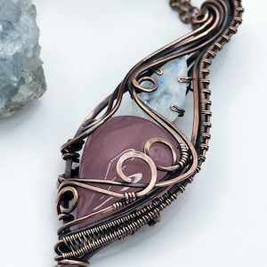 Può includere: Ciondolo fatto a mano con una gemma rosa a forma di cuore lucidata e una pietra bianca più piccola. Le pietre sono racchiuse in un intricato filo di rame, sospese a una catena abbinata. Il design del ciondolo è organico e artistico.