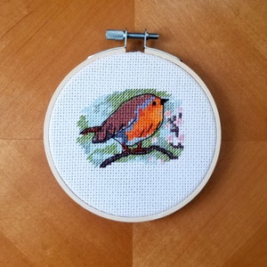 以下が含まれることがあります： ピンクの花が咲いた枝に止まっているコマドリのクロスステッチ刺繍。コマドリは茶色、オレンジ、白です。刺繍は丸い木製の枠で囲まれています。