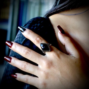 May include: A black gemstone ring with a geometric design. The ring features a large, dark, square-cut stone set in a decorative metal band. The ring is on a hand with dark red nail polish.